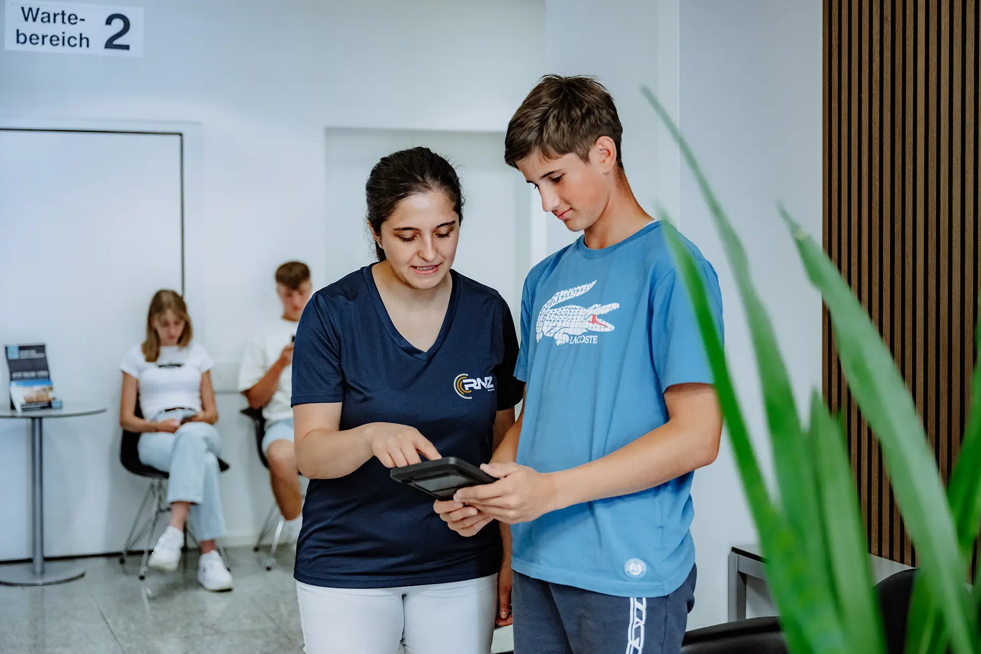Medizinische Fachangestellte unterstützt einen jungen Patienten bei der Anmeldung im Wartebereich.